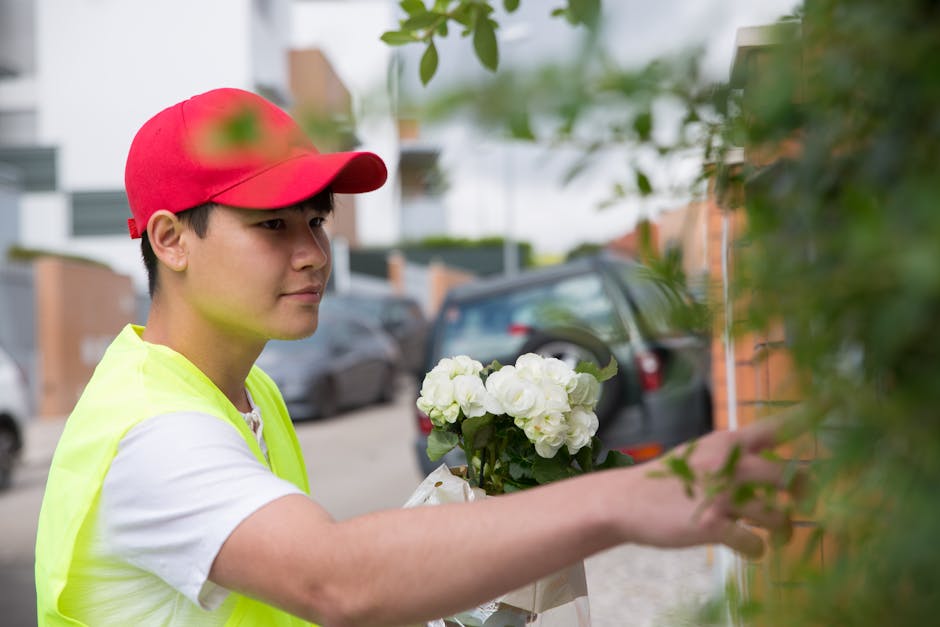 A young male delivery worker wearing a red cap and a high-visibility vest is holding a bouquet of fresh white flowers, including roses and hydrangeas, wrapped in clear cellophane. He is reaching out t