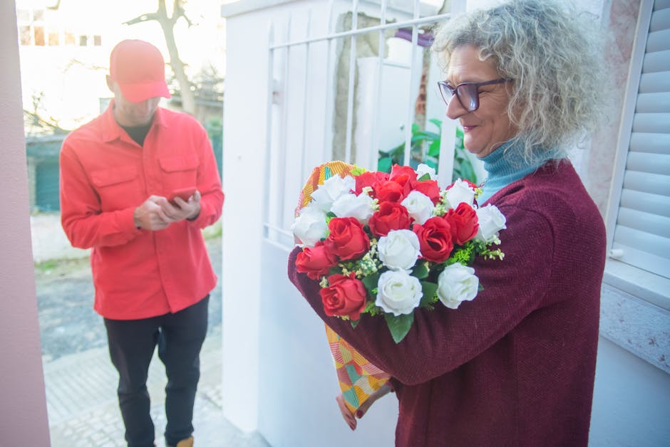 A woman with curly gray hair, wearing glasses, a blue turtleneck, and a burgundy sweater, holds a vibrant floral gift bouquet featuring red and white roses, wrapped in colorful paper. She is standing 