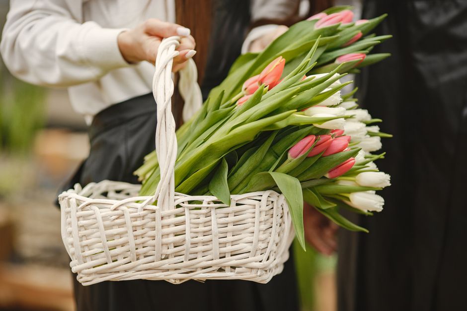 A person holding a white wicker basket filled with fresh tulips in shades of pink and white, with green stems and leaves visible. The basket is wrapped with a white handle, and the flowers are arrange