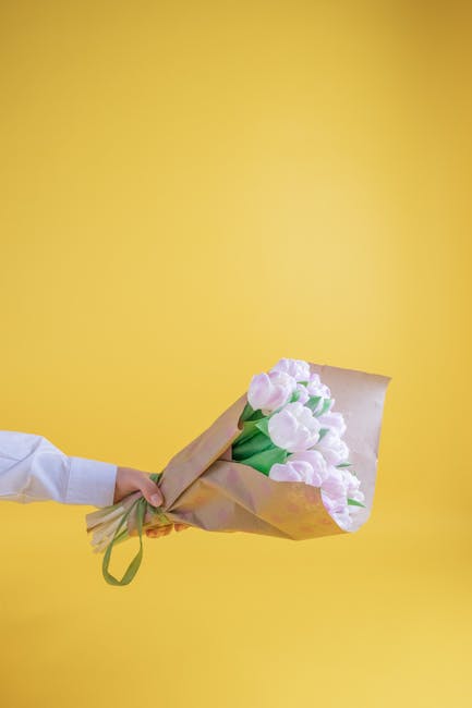A person’s hand is visible, extending from the left side of the image, holding a bouquet of fresh, pale pink tulips wrapped in light brown kraft paper with a green ribbon. The tulips have smooth, sl