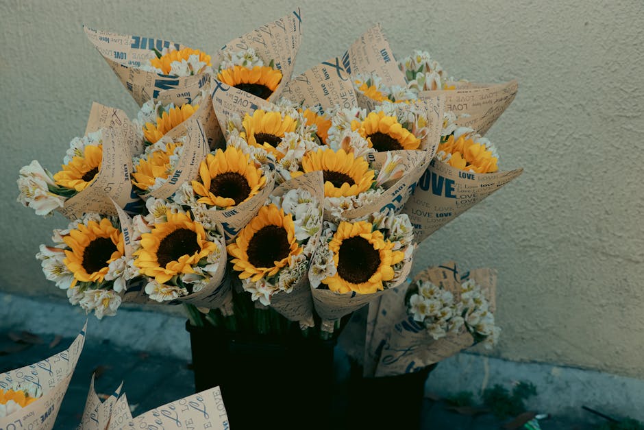 A bouquet of fresh sunflowers with bright yellow petals and dark centers, wrapped in light brown patterned paper with words like 'LOVE' and 'JOY' in blue, sits in a black container against a textured 